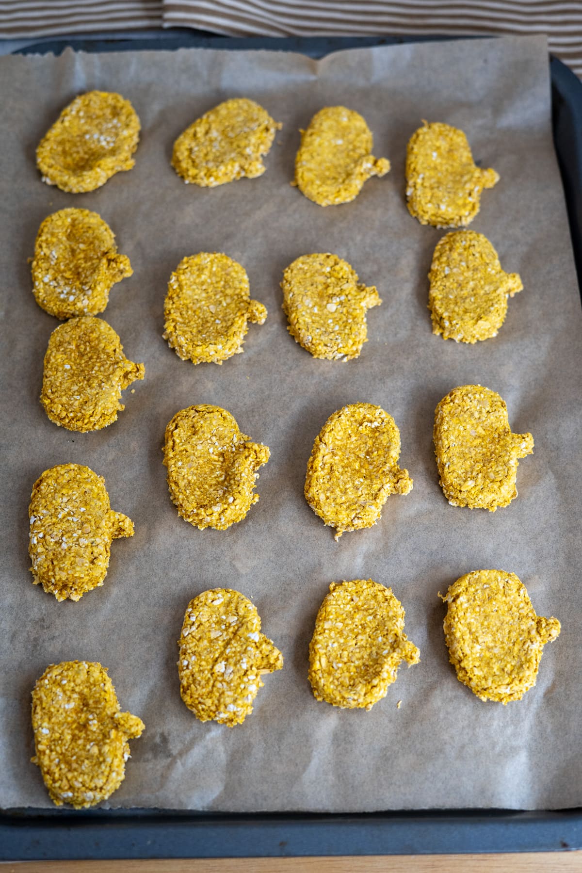 A baking tray lined with greaseproof paper holds fifteen uncooked, breaded goat's milk pumpkin dog biscuits arranged in rows.