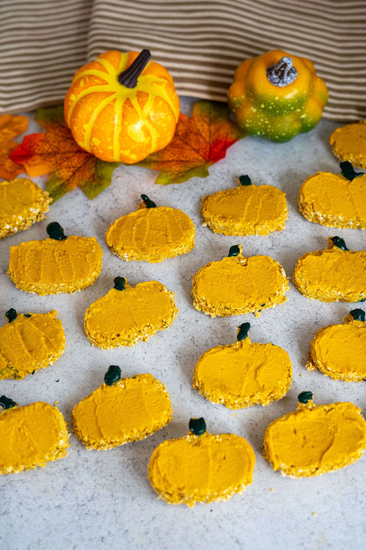 Pumpkin-shaped biscuits with orange icing and green stalks are arranged on a surface, accompanied by goat milk pumpkin dog biscuits, with artificial pumpkins and autumn leaves in the background.
