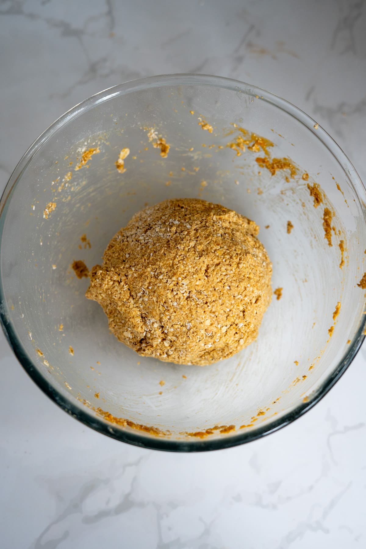 A ball of raw dough studded with visible oats and enriched for goat milk pumpkin dog biscuits sits in a glass mixing bowl on a white marble worktop.