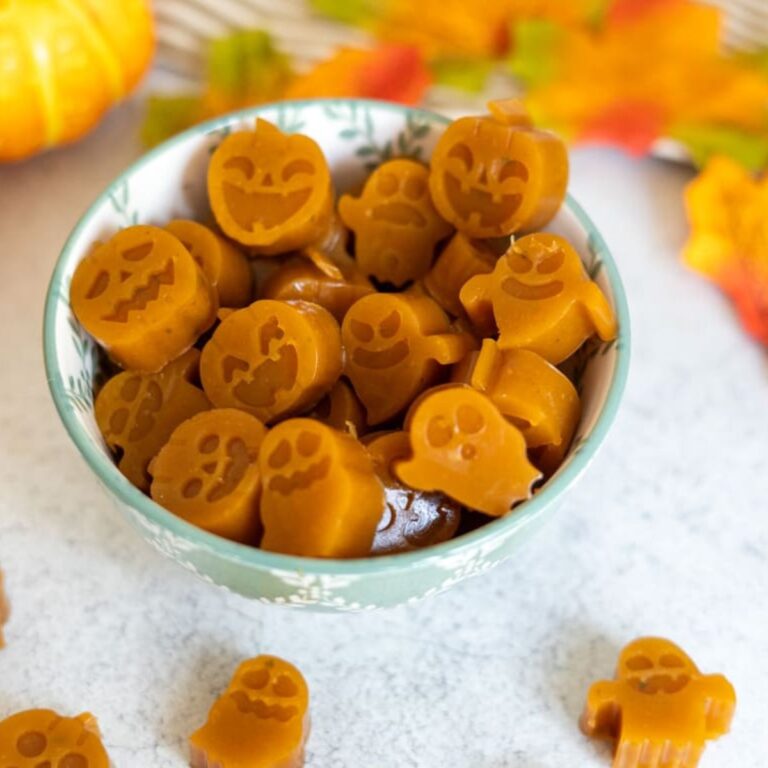 A bowl filled with pumpkin-shaped and ghost-shaped orange sweets sits on a light surface with autumn leaves in the background.
