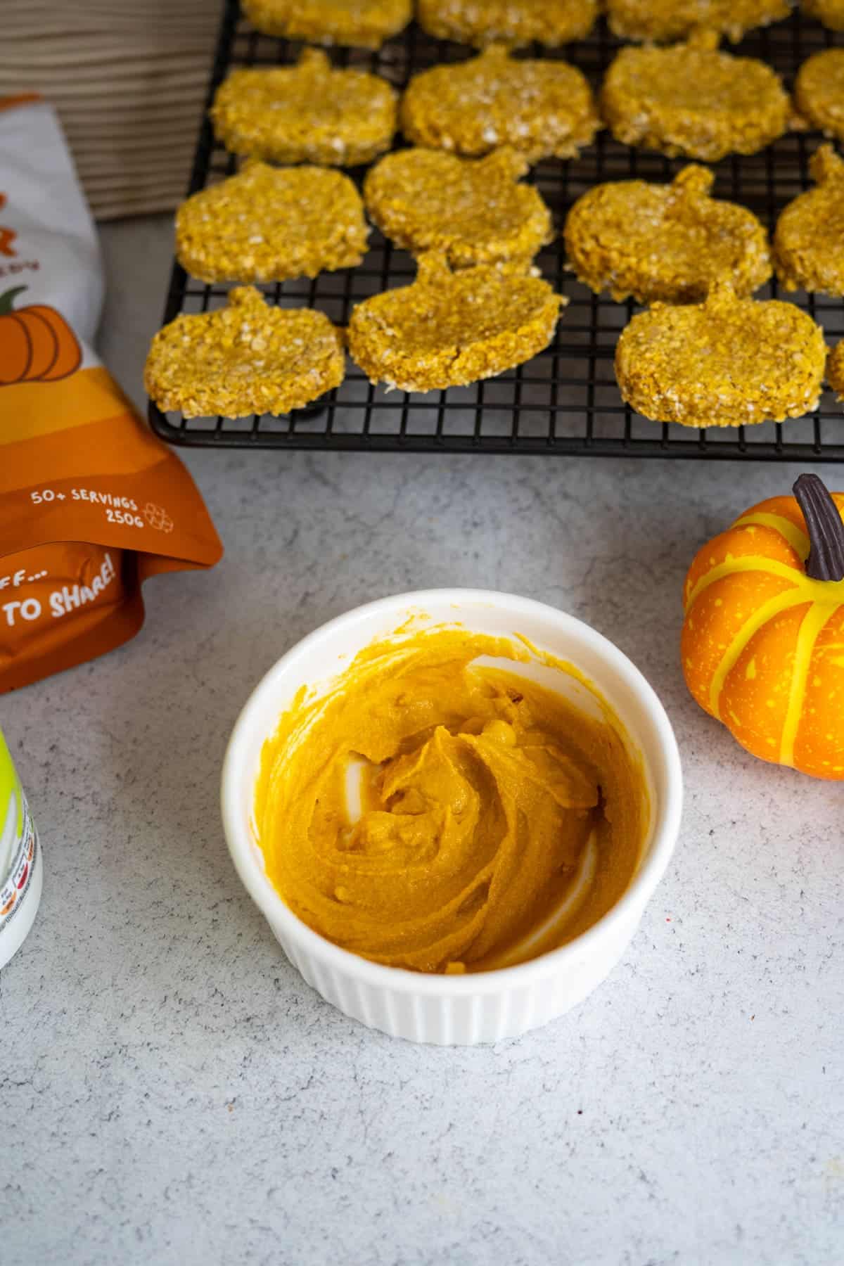 A bowl of orange batter sits on a worktop next to a small pumpkin, hinting at the homemade goodness of goat milk pumpkin dog treats, with round breaded patties cooling on a wire rack in the background.