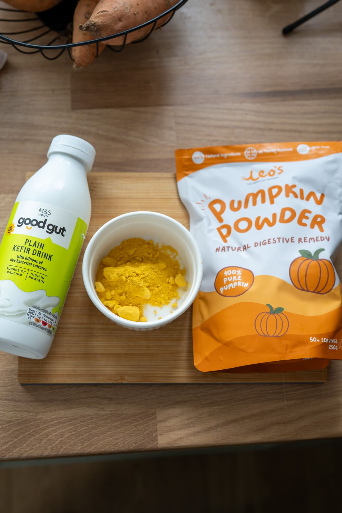 A bottle of plain kefir drink, a bowl of yellow powder, and a bag of pumpkin powder are arranged on a wooden surface, alongside some goat milk pumpkin dog biscuits for a nutritious treat.