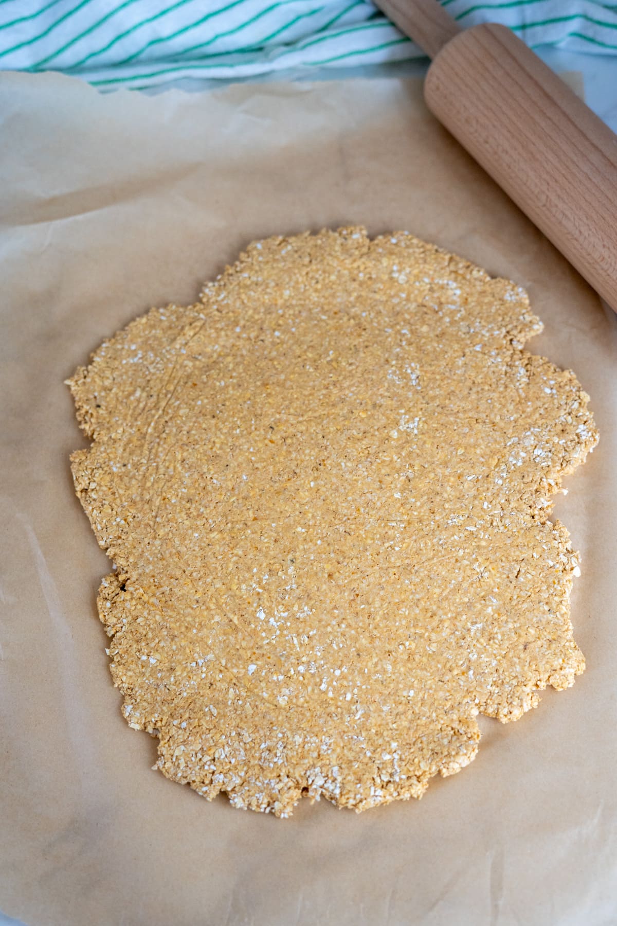 A rolled-out, uneven round of dough for goat milk pumpkin dog biscuits rests on baking parchment with a wooden rolling pin nearby, all set atop a striped cloth background.