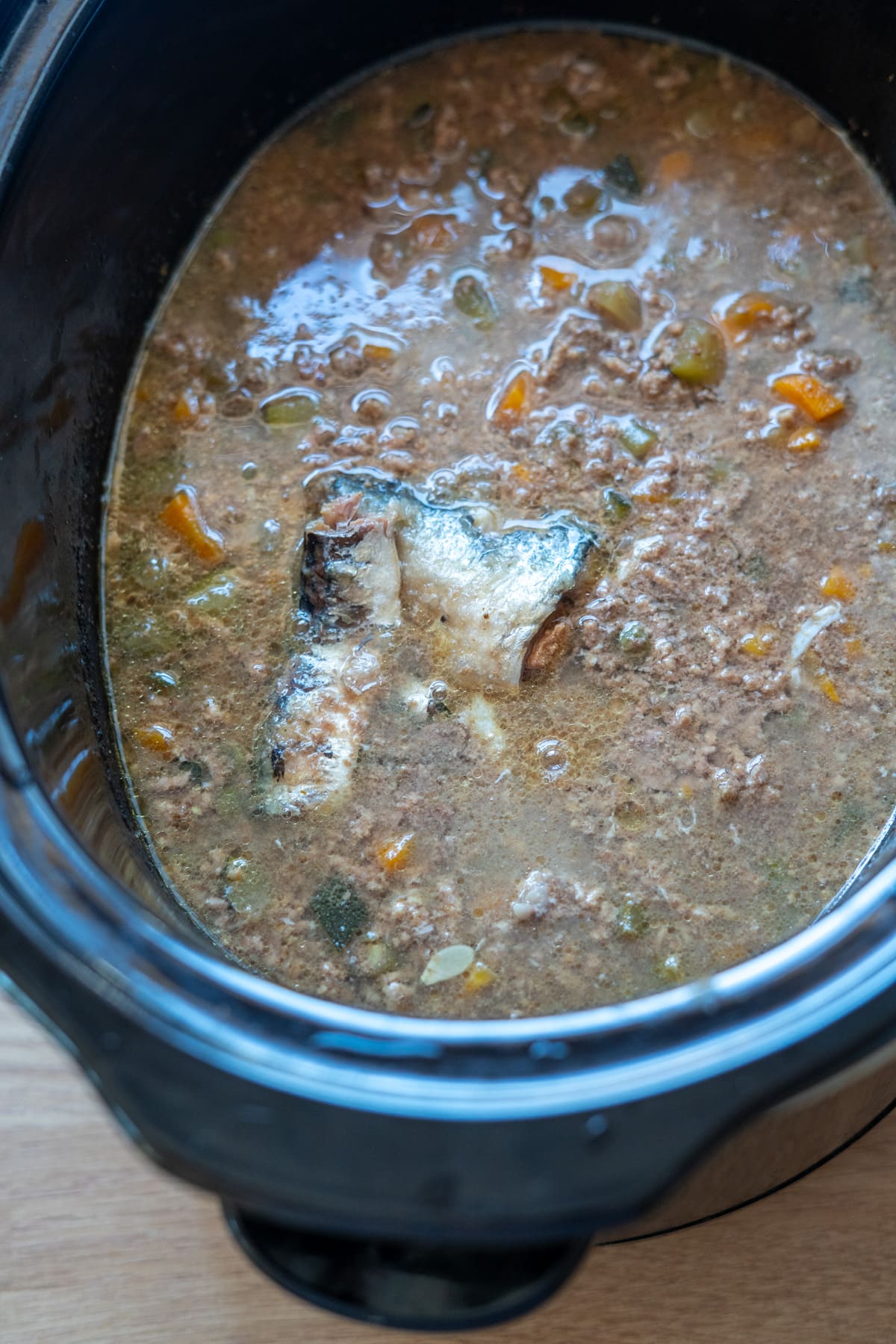 A close-up of a slow cooker filled with a chunky Slow Cooker Beef Dog Food stew, featuring minced meat, vegetables, and two fish pieces simmering in savoury broth.