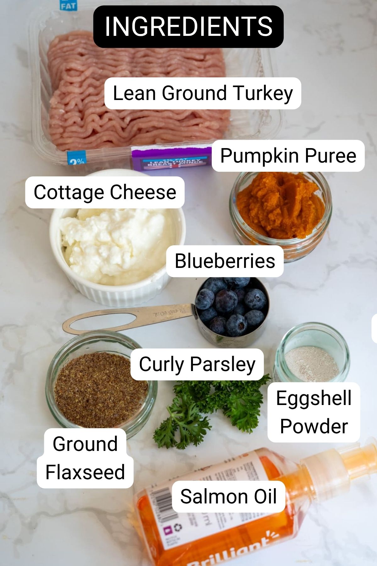 An assortment of labelled ingredients on a worktop, including lean minced turkey, cottage cheese, pumpkin purée, blueberries, curly parsley, flaxseed, eggshell powder, and salmon oil.