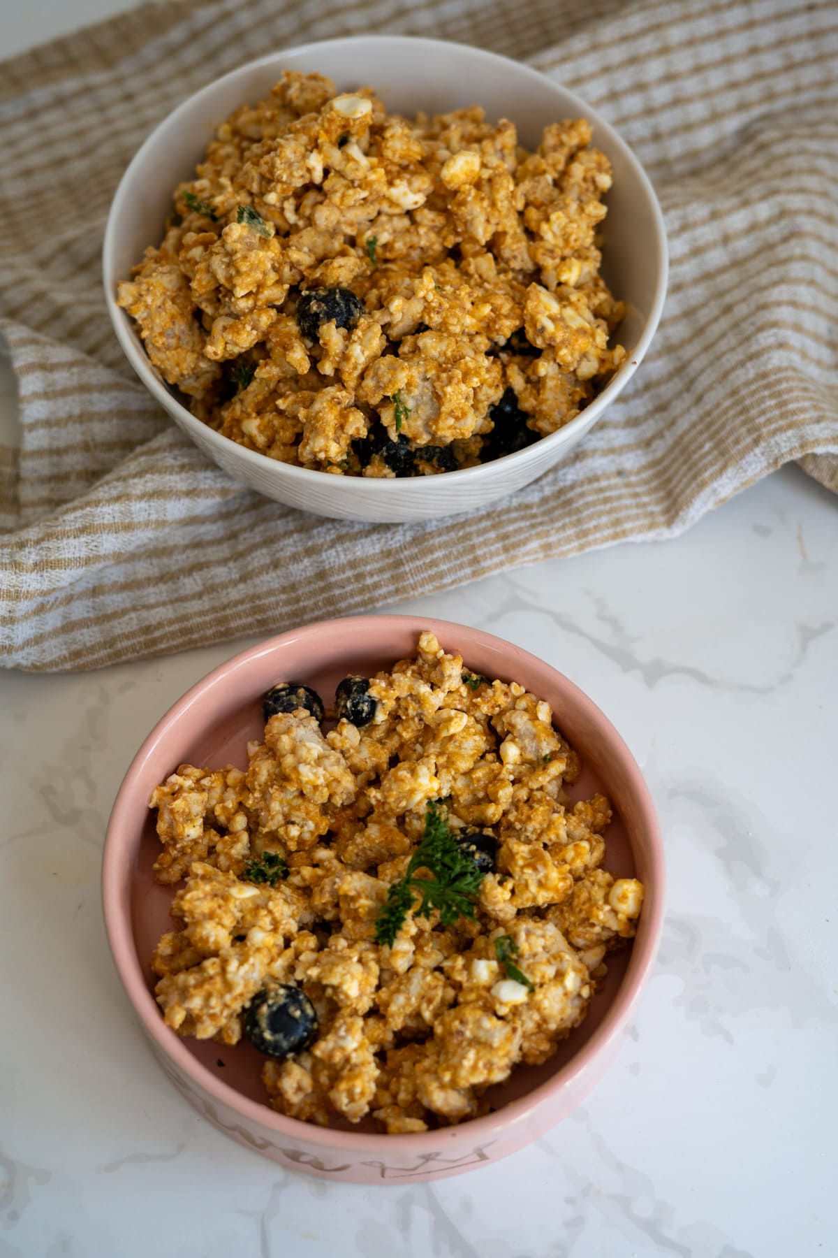 Two bowls of scrambled tofu with herbs and black olives sit on a striped cloth and marble worktop, offering a colourful plant-based twist that contrasts with a traditional turkey breakfast for dogs.