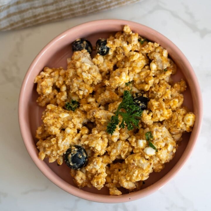 A pink bowl filled with a mixture of scrambled eggs, olives, cottage cheese, and seasoning, garnished with a sprig of parsley.