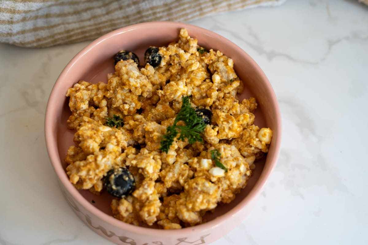 A pink bowl filled with a mixture of crumbled paneer, black olives, and spices, garnished with fresh herbs on a marble surface.