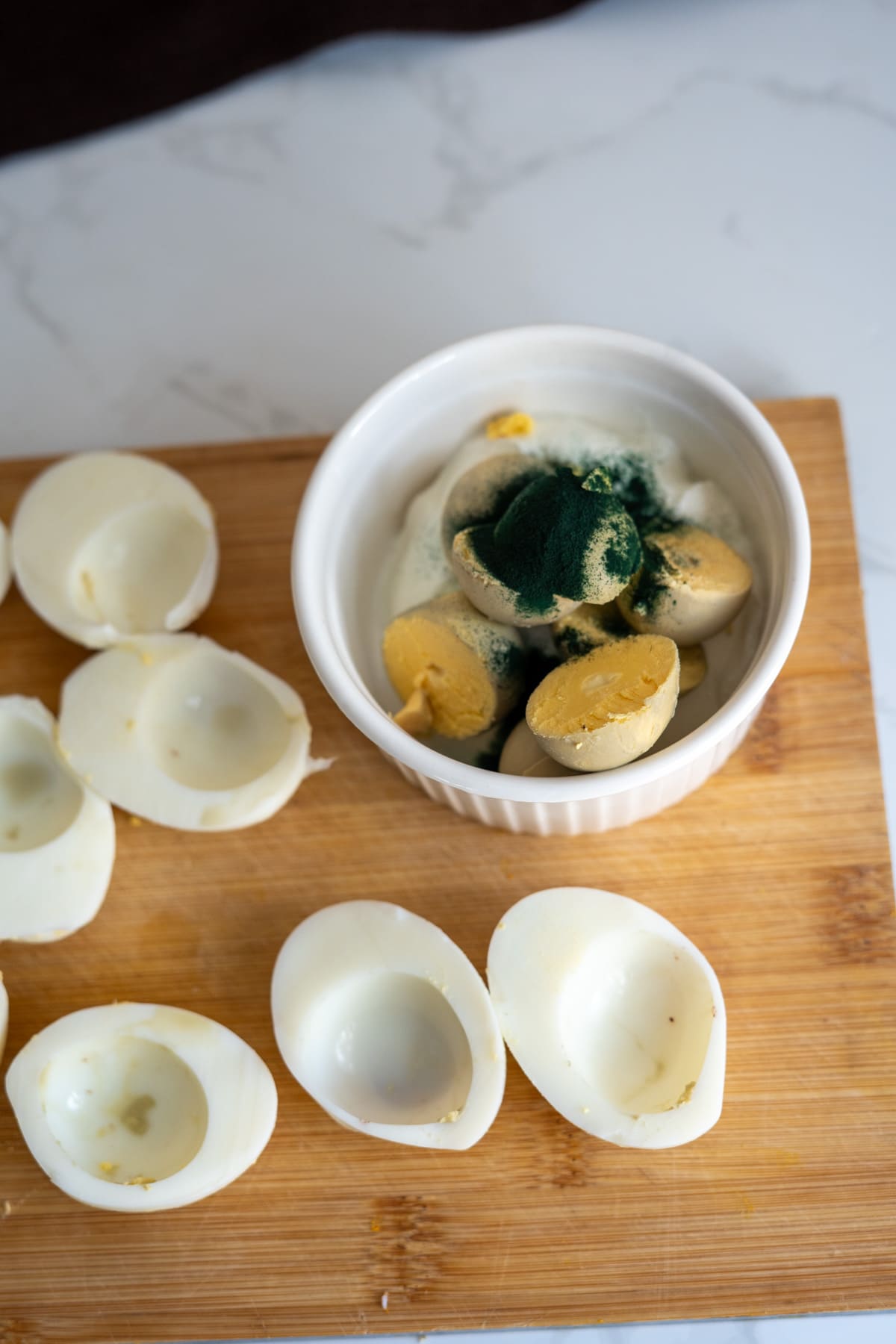 Hard-boiled egg whites on a wooden chopping board next to a bowl of yolks mixed with green powder, prepared as Halloween devilled eggs for dogs.