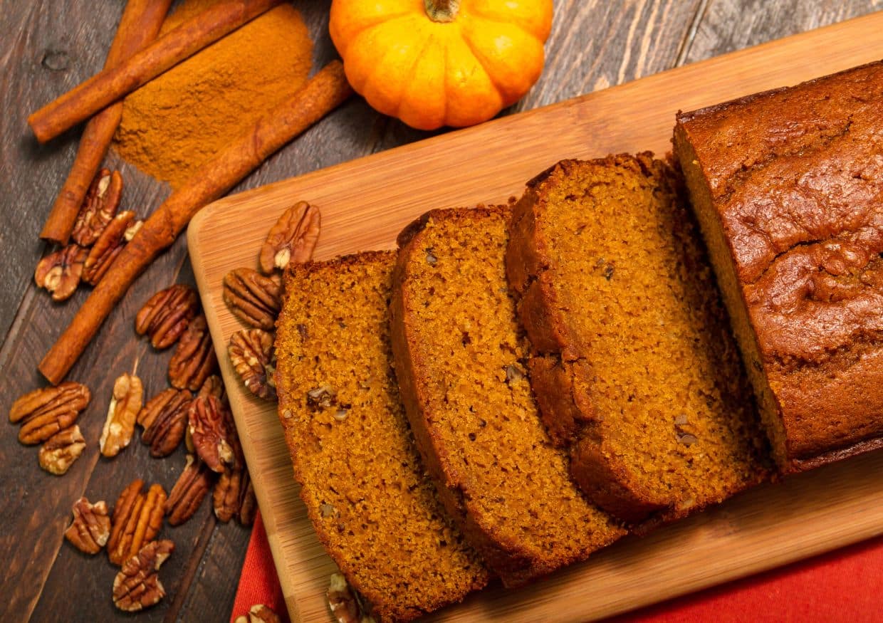 Sliced pumpkin bread on a wooden board with pecans, cinnamon sticks, ground cinnamon, and a small pumpkin on a rustic table.