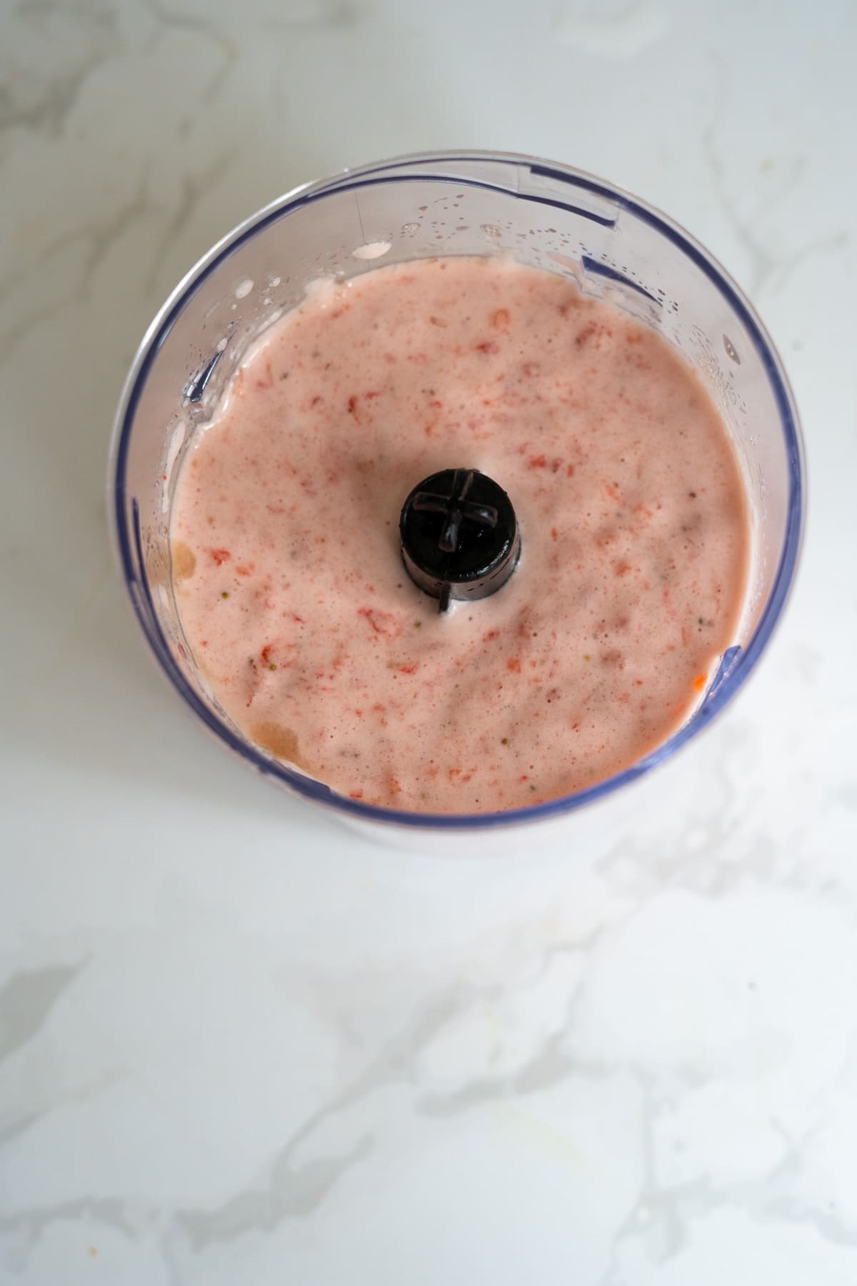 A food processor bowl filled with a blended pink mixture, perfect for making strawberry gummies for dogs, sits on a white marble surface.