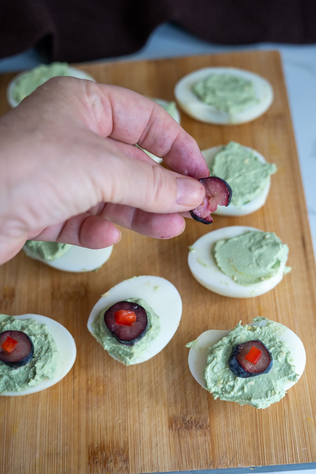 A hand places a sliced grape topped with red pepper onto a Halloween devilled egg for dogs, filled with green herb spread on a wooden board.