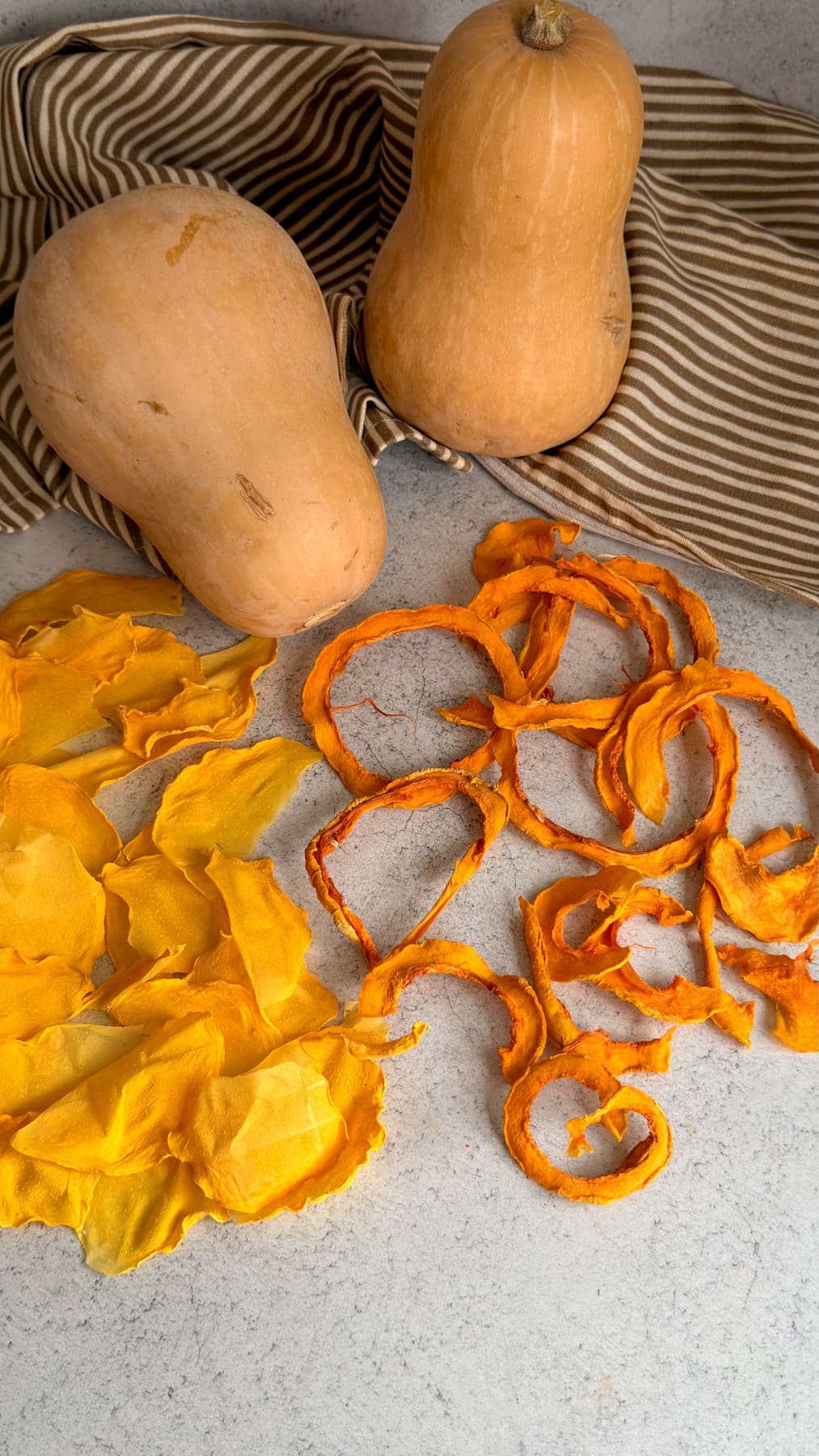 Two whole butternut squash next to piles of thin, dried butternut squash crisps for dogs on a light-coloured surface.