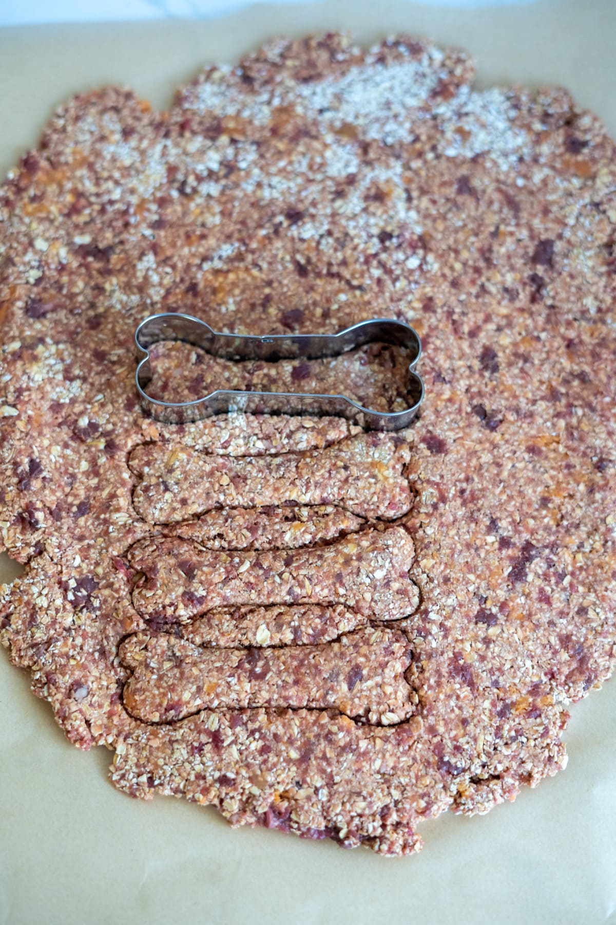A bone-shaped biscuit cutter rests on a rolled-out sheet of lamb and sweet potato dog treat dough, with several bone-shaped treats already cut out.