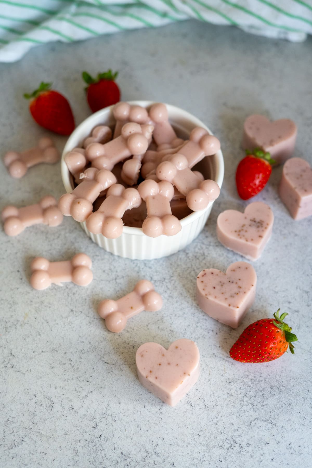 A white bowl filled with pink, bone-shaped strawberry gummies for dogs sits on a worktop, surrounded by more bone and heart-shaped treats and several whole strawberries.