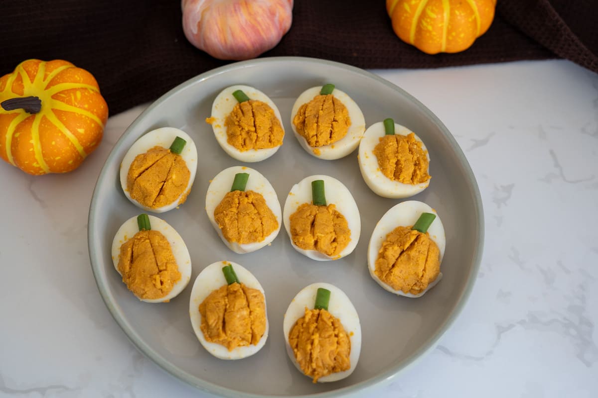 A plate of pumpkin devilled eggs for dogs with orange-dyed yolks shaped like pumpkins, each topped with a chive stem and surrounded by decorative mini pumpkins.