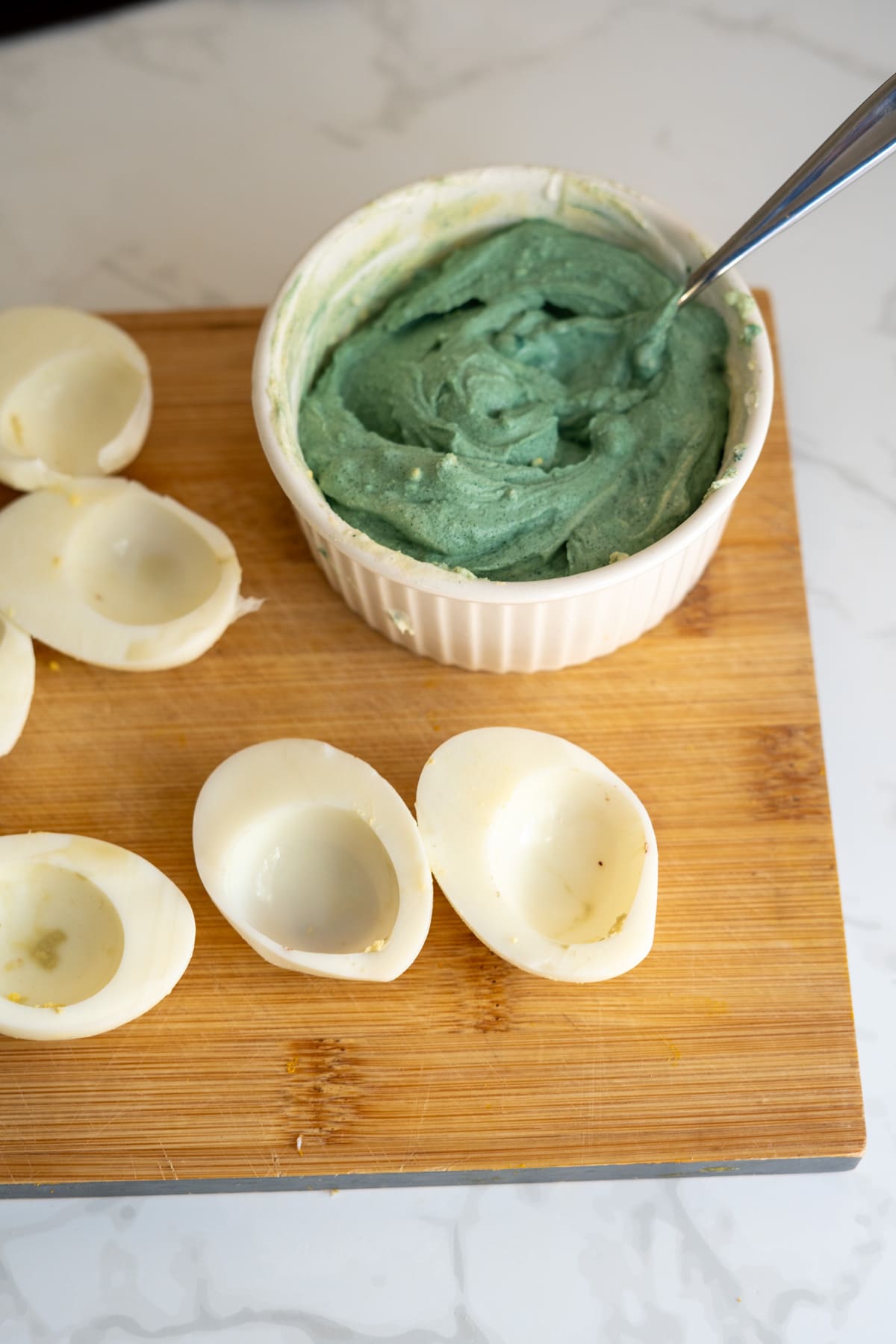 A bowl of blue filling for Hallowe'en devilled eggs for dogs next to several halved hard-boiled egg whites on a wooden chopping board.
