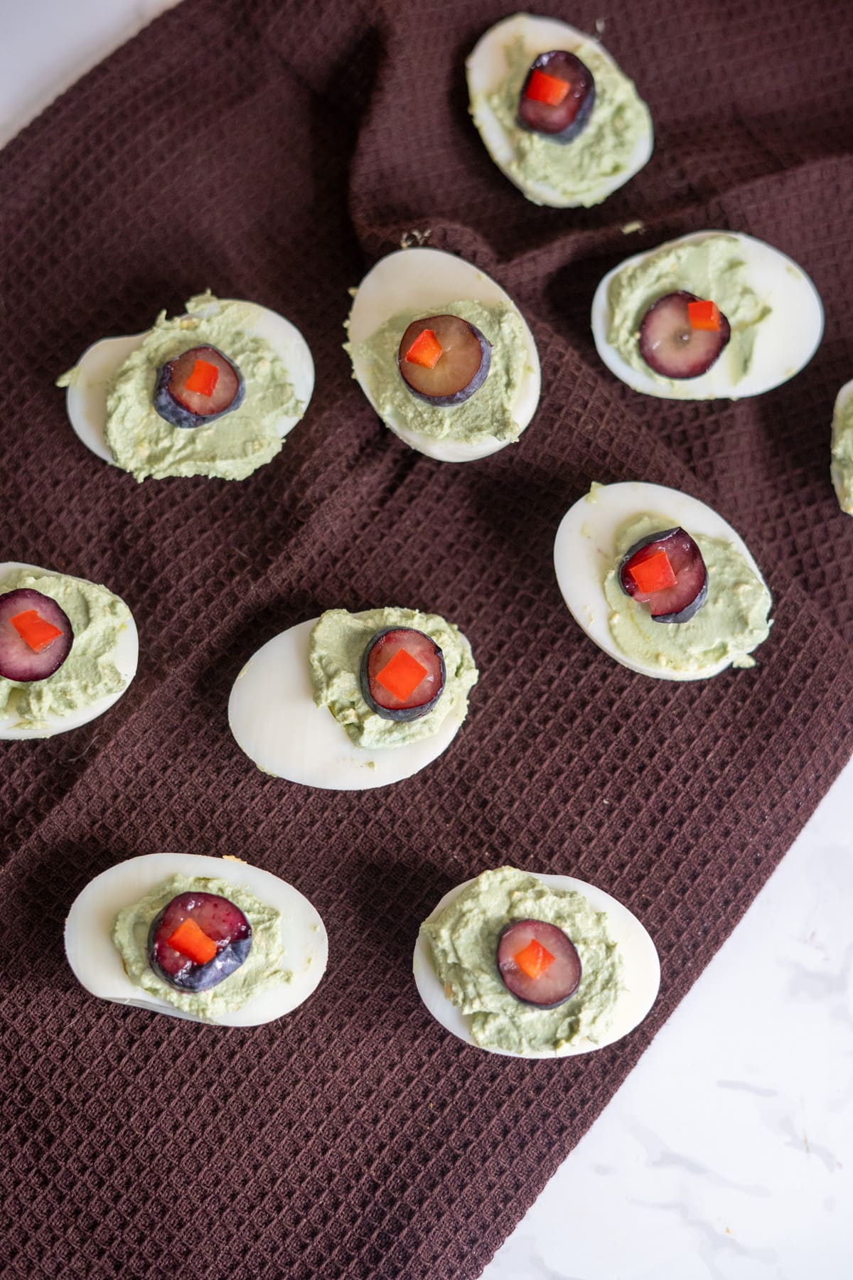 Hallowe’en devilled eggs for dogs, topped with green filling, a grape slice, and a piece of red pepper, arranged on a brown textured cloth.