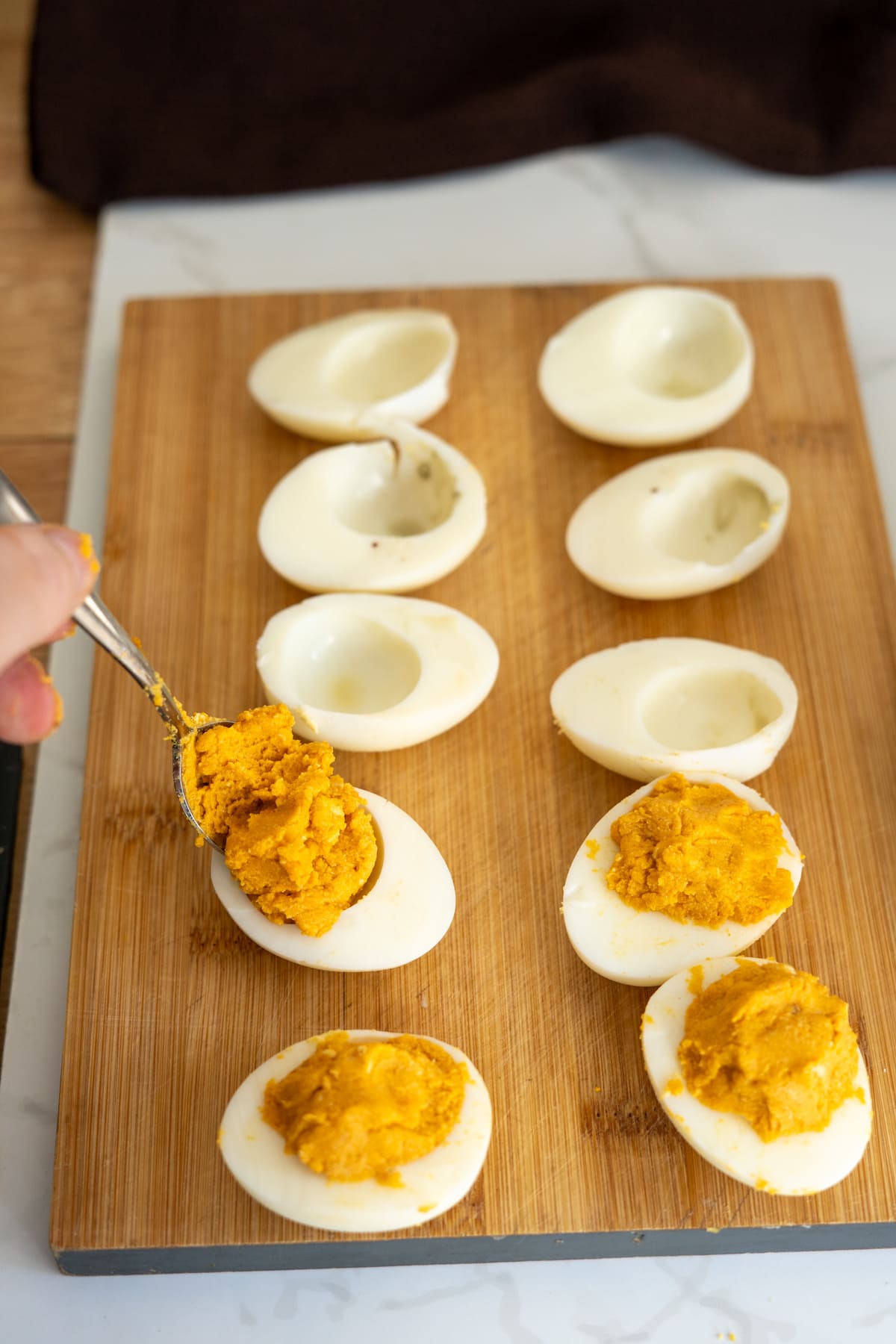 A hand uses a spoon to fill hollowed-out hard-boiled egg whites with a yellow yolk mixture on a wooden chopping board to make pumpkin devilled eggs for dogs.