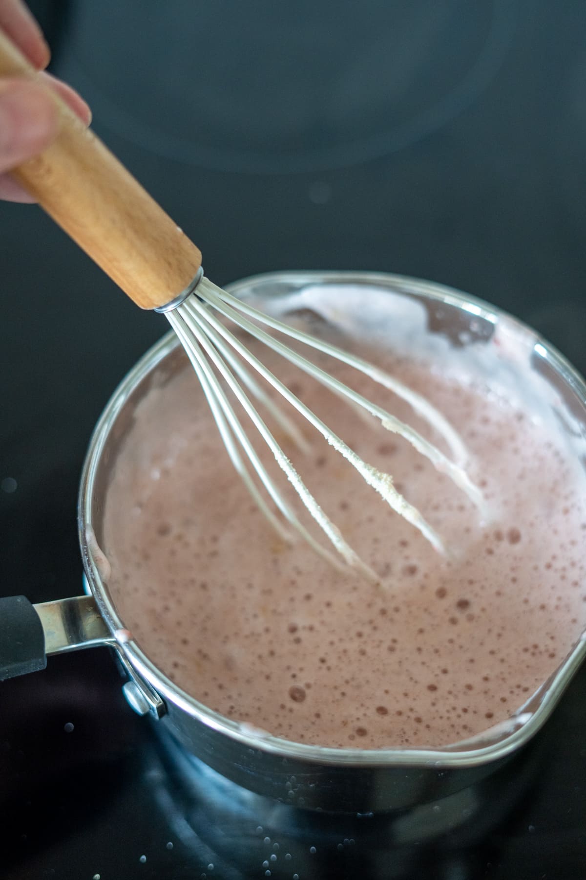 A hand holds a whisk over a saucepan of frothy, light brown liquid—perhaps mixing up homemade strawberry sweets for dogs on the hob.