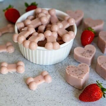 A white bowl filled with pink bone-shaped treats, surrounded by heart-shaped treats and strawberries on a light surface.