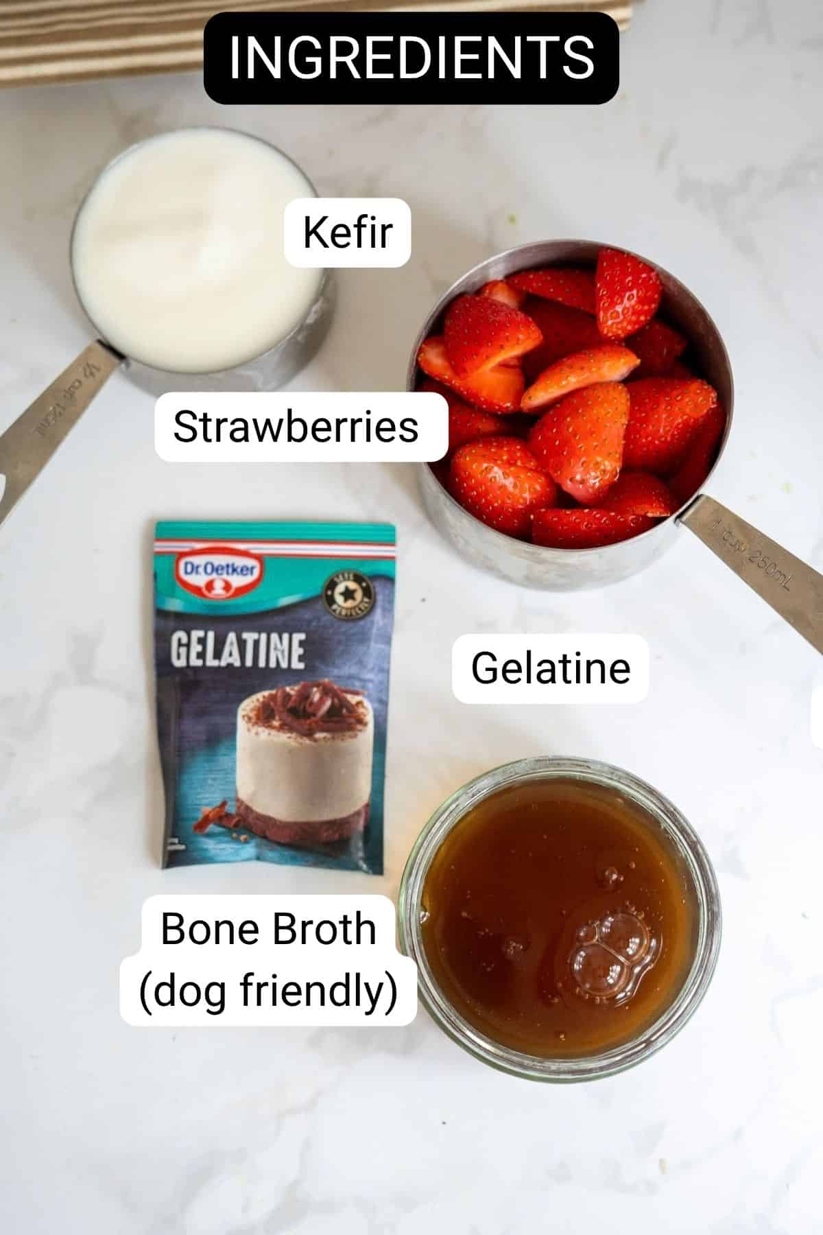 Measuring jugs with kefir, strawberries, a packet of gelatine, and a jar of dog-friendly bone broth for making strawberry gummies for dogs, all arranged on a white surface.