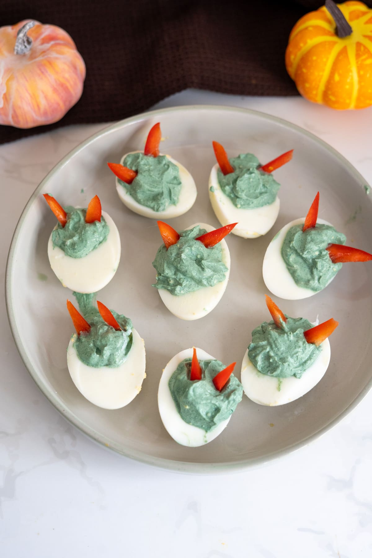 Halloween devilled eggs for dogs, featuring green-tinted filling and red pepper horns, are arranged on a plate and surrounded by two small decorative pumpkins.