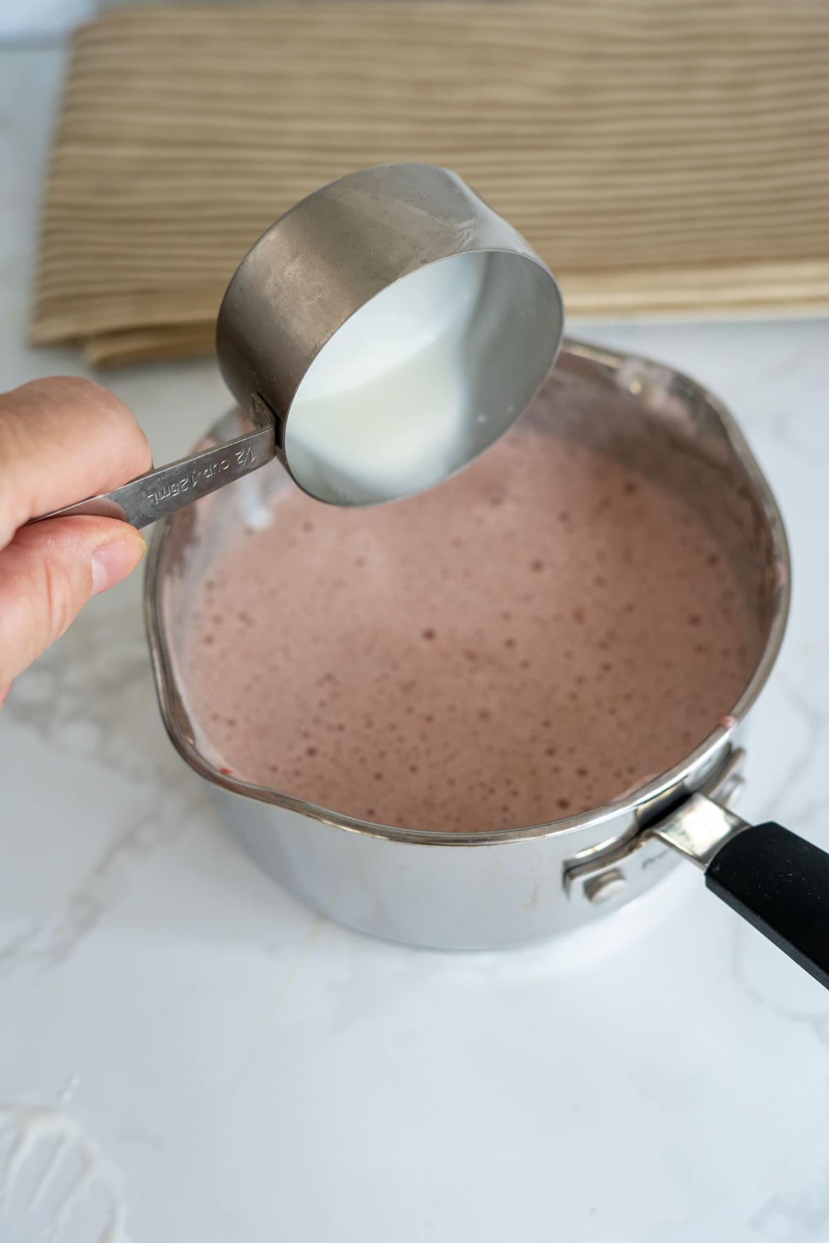 A hand pours milk from a metal measuring jug into a saucepan filled with a frothy pink liquid—perfect for making strawberry gummies for dogs—on a white worktop.