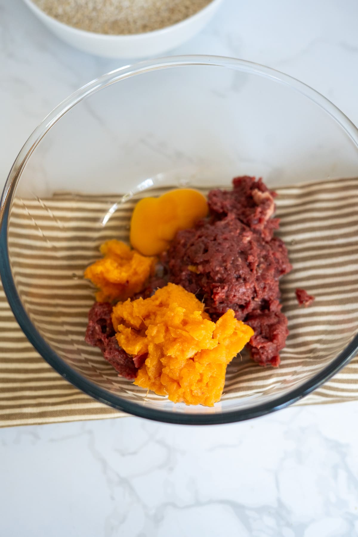A glass bowl containing raw minced lamb and mashed pumpkin for sweet potato dog treats, with a raw egg yolk.