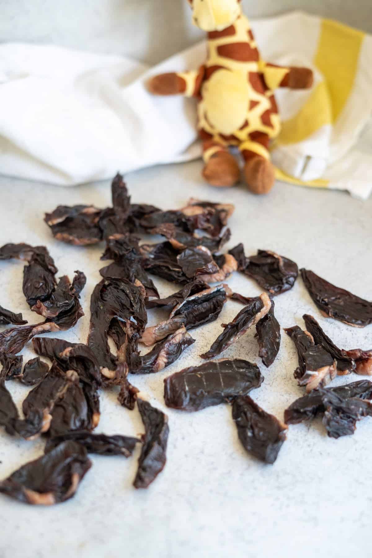 A pile of dark, dried lamb heart dog treats with a small plush giraffe in the background.
