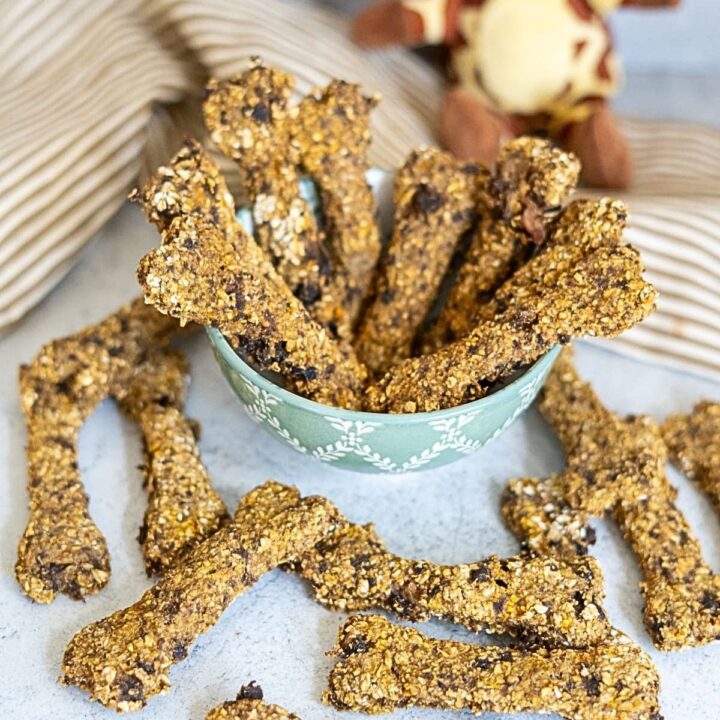 Bone-shaped dog treats piled in a small green bowl and scattered on a white surface.