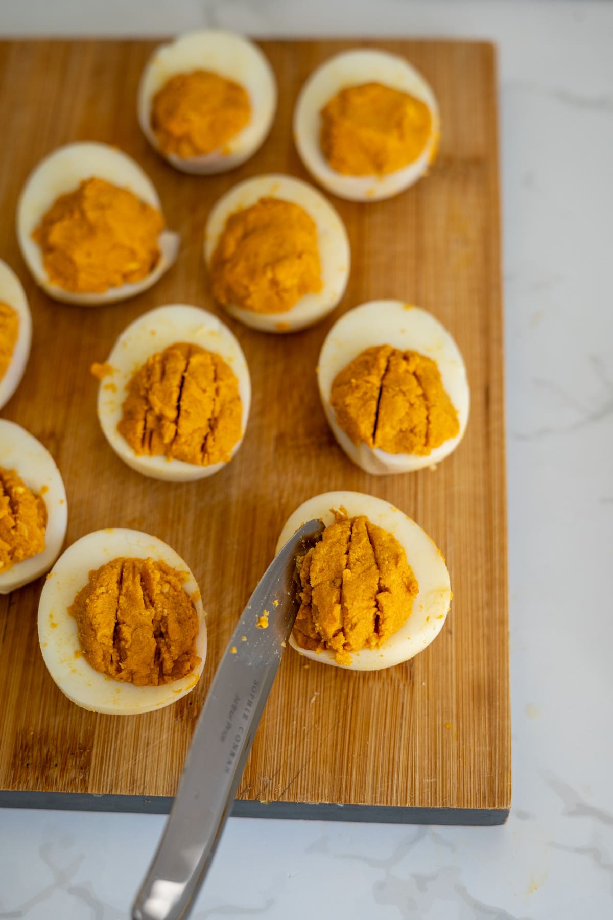 A knife spreads orange pumpkin devilled eggs for dogs filling into halved hard-boiled eggs arranged on a wooden chopping board.