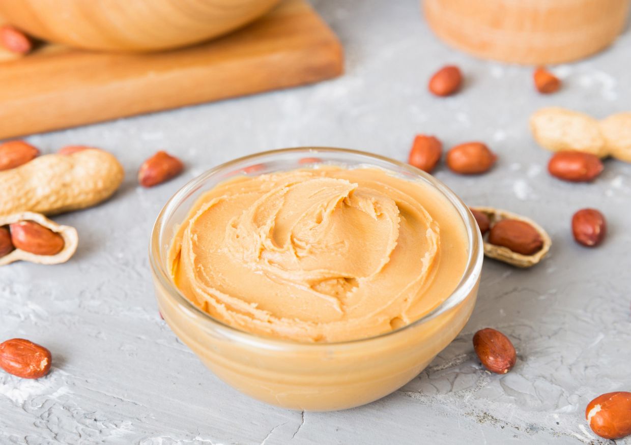 A glass bowl of smooth peanut butter, which some owners ask if dogs can eat, surrounded by shelled and unshelled peanuts.