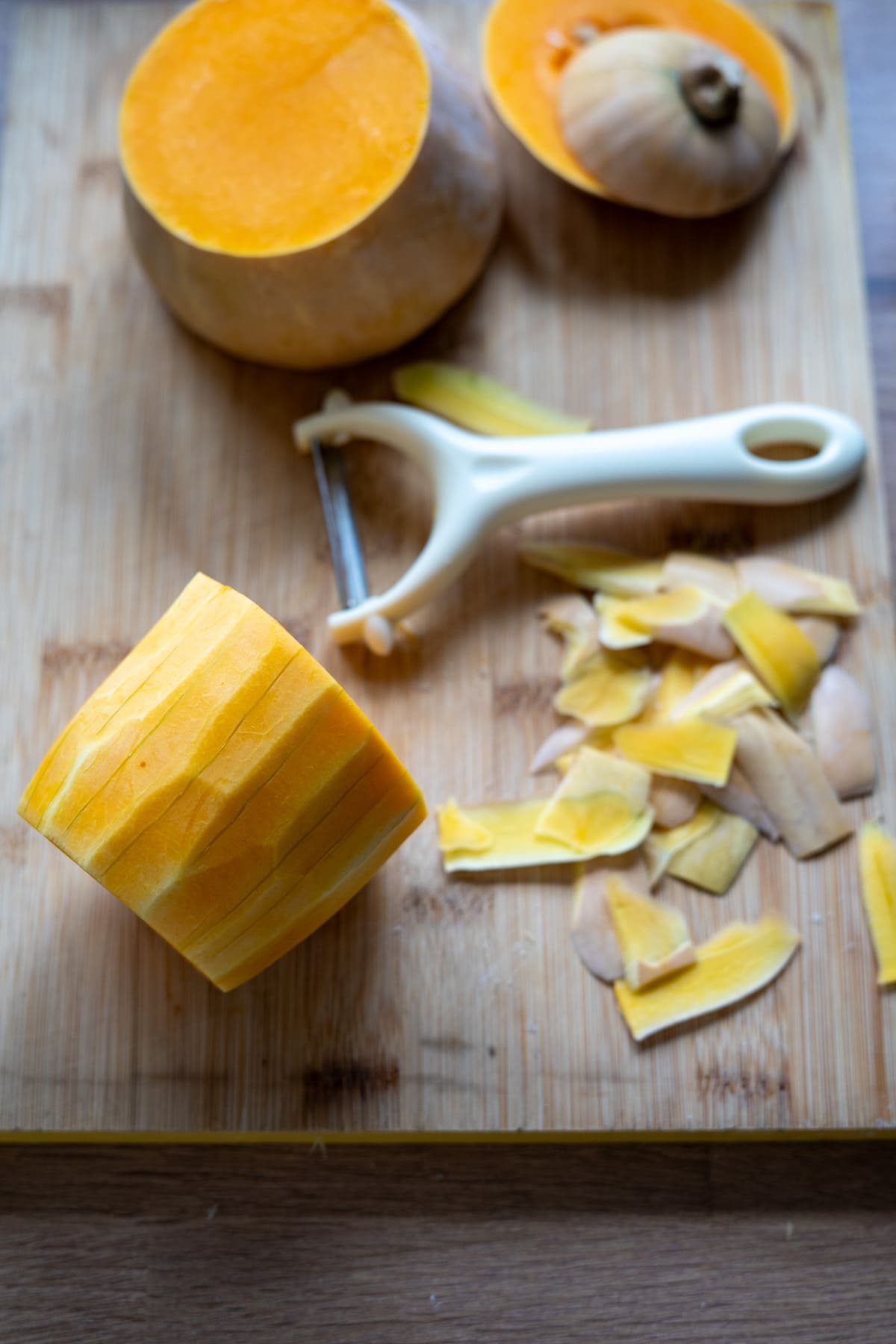 A peeled butternut squash for making dog crisps, a vegetable peeler, and squash peelings on a wooden chopping board.