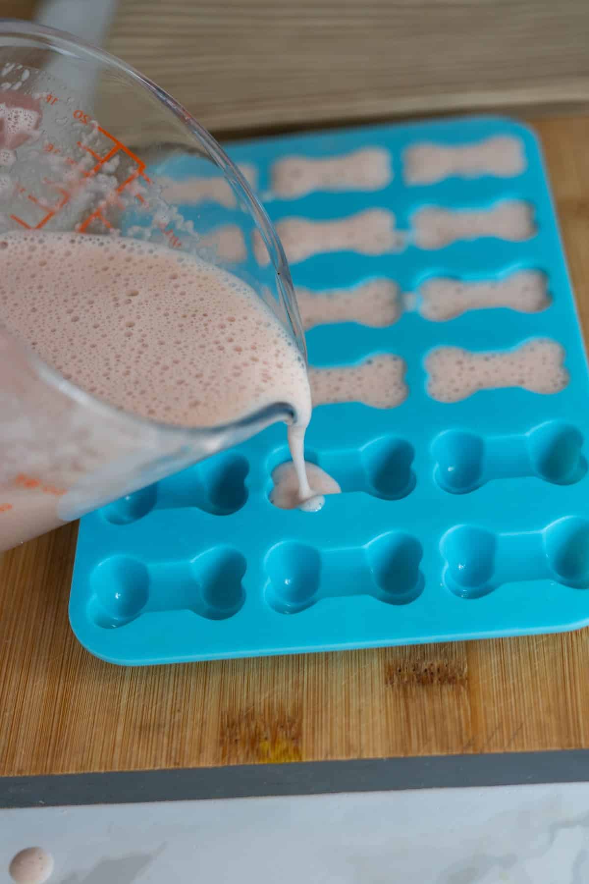 A measuring jug pours a pink strawberry gummies for dogs mixture into a blue silicone mould shaped like dog bones, on a wooden surface.