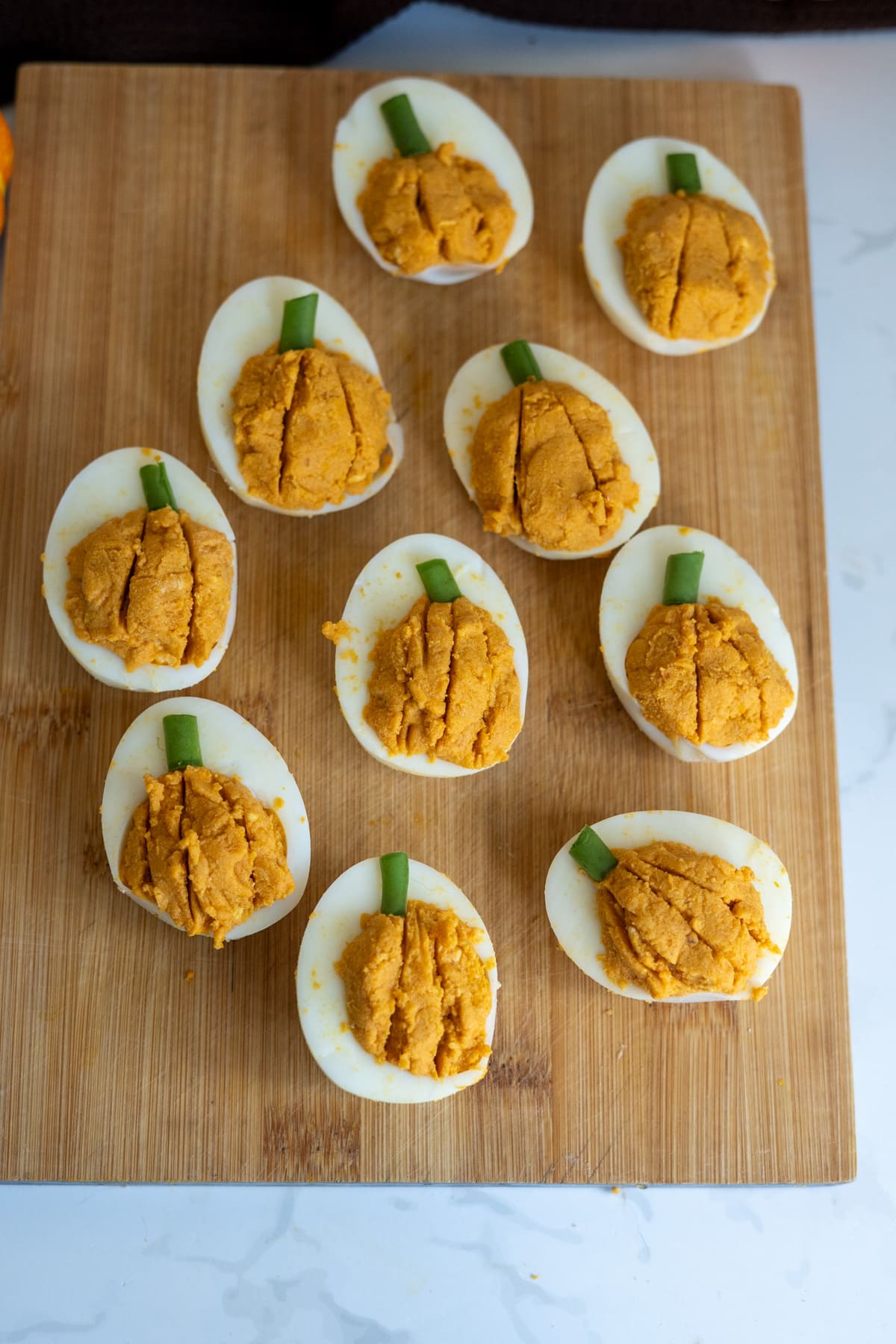 Pumpkin devilled eggs for dogs arranged on a wooden chopping board, featuring orange-tinted filling shaped like pumpkins and topped with spring onion stems.