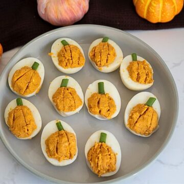 Nine devilled eggs arranged on a grey plate, with the yolk filling shaped like pumpkins and topped with small pieces of green onion stems.