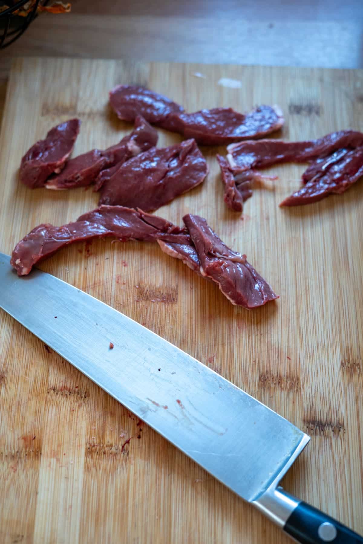 Raw sliced lamb heart for dog treats on a wooden chopping board next to a chef’s knife.