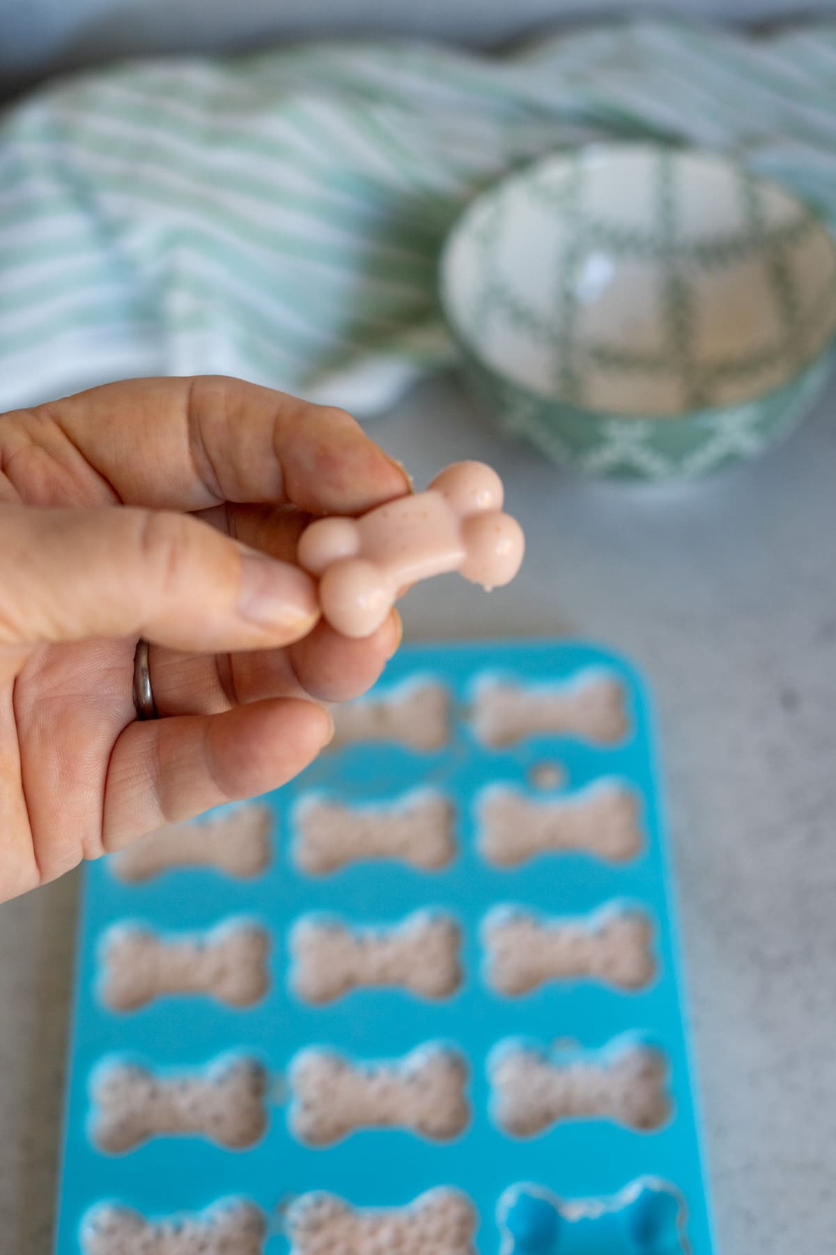 A hand holds a small, bone-shaped strawberry gummy for dogs above a blue silicone mould filled with similar treats. A bowl and striped cloth are in the background.