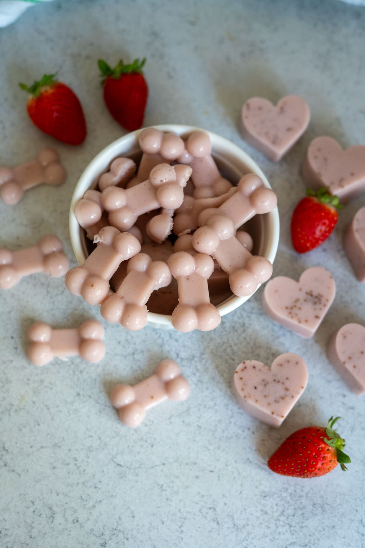 A bowl of pink, bone-shaped strawberry gums for dogs surrounded by more bone- and heart-shaped treats, with several whole strawberries on a light surface.