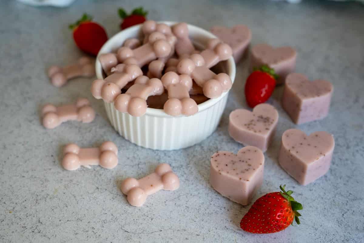 A white bowl filled with pink dog bone-shaped strawberry gummies for dogs sits on a counter, surrounded by more bone and heart-shaped treats and fresh strawberries.
