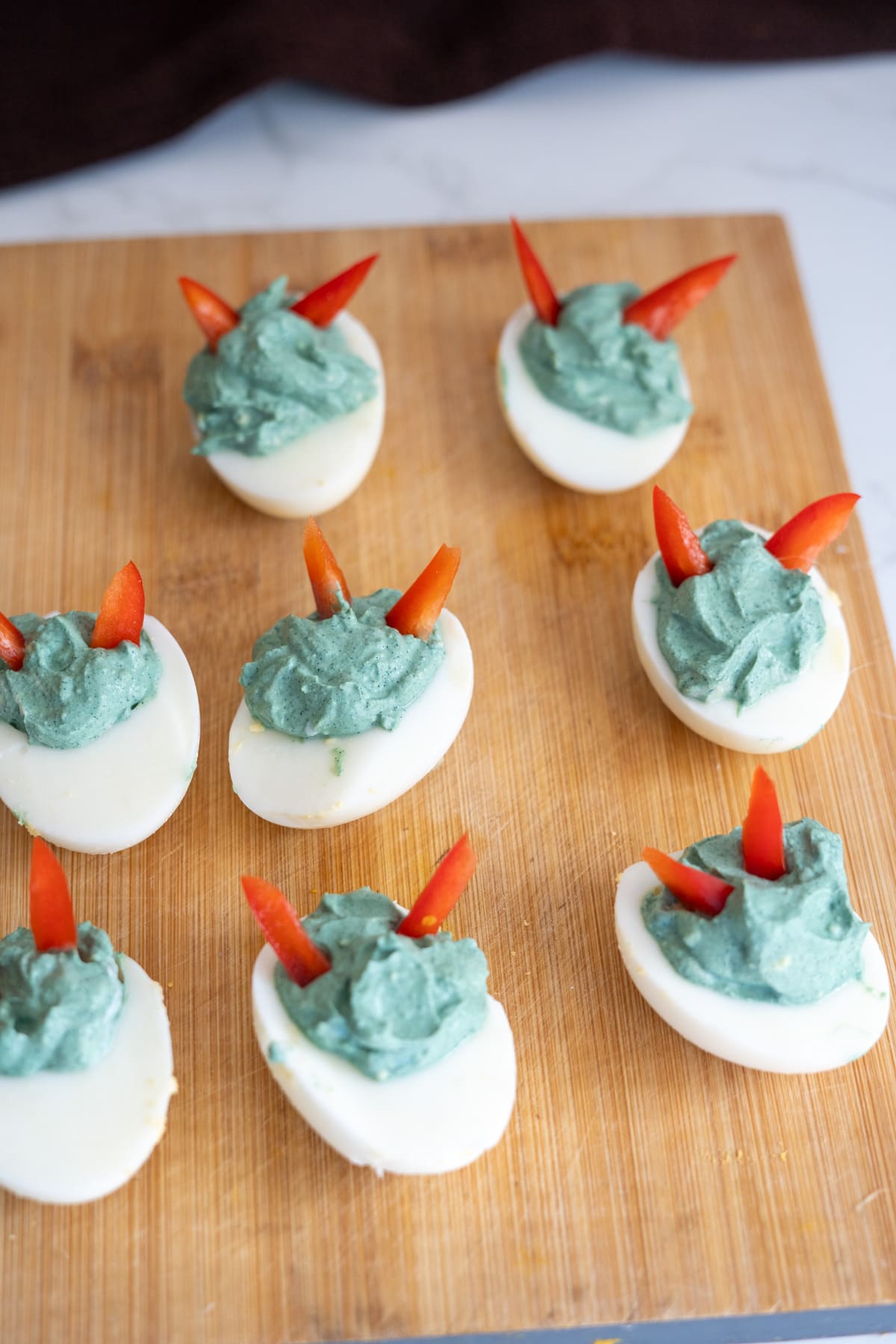 Halloween devilled eggs for dogs, filled with blue-tinted yolk and topped with red pepper slices arranged on a wooden board.
